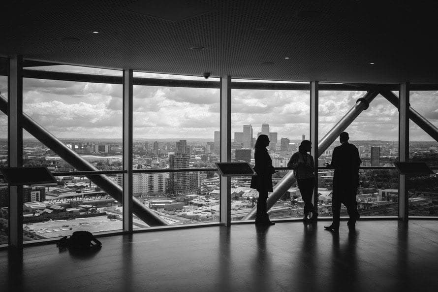 silhouetted figures converse by large windows, overlooking a vibrant urban landscape in a modern building.