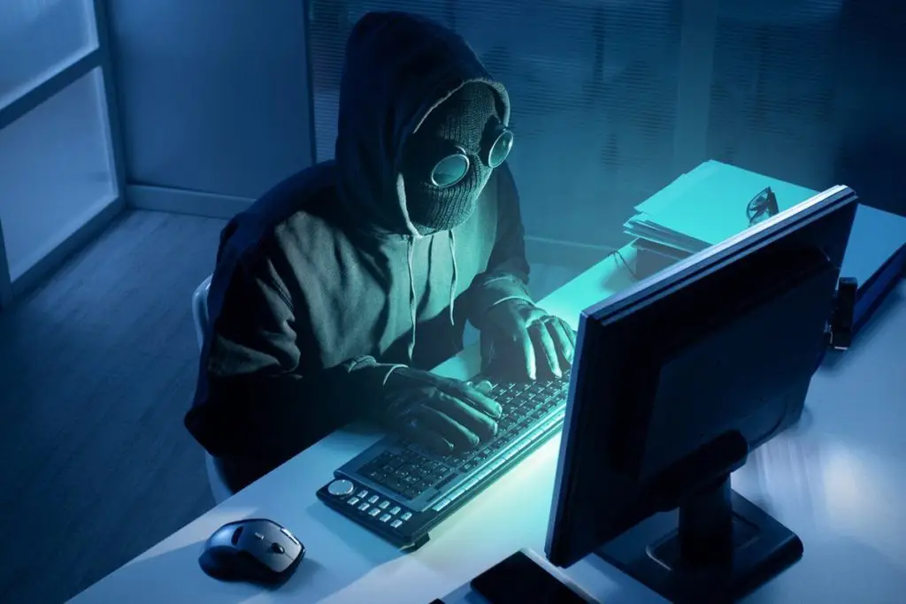 Three New Threats To Look Out For In The Coming Year 1 hooded hacker immersed in digital secrets, illuminated by a blue-glowing computer screen.
