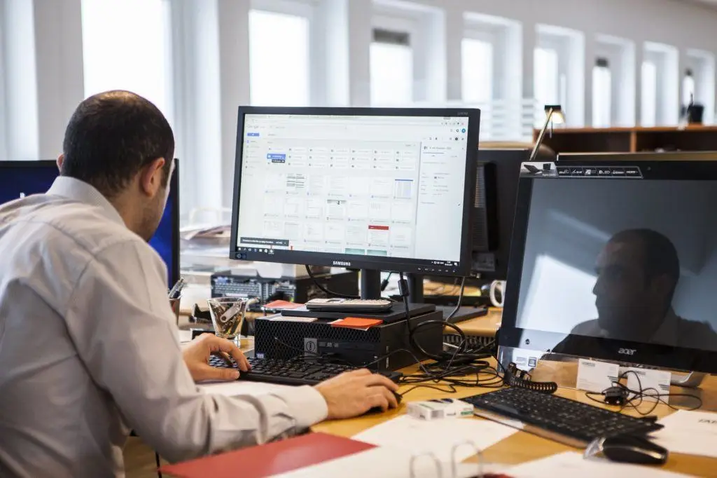 man working at a tidy desk with dual monitors in a bright, modern office.
