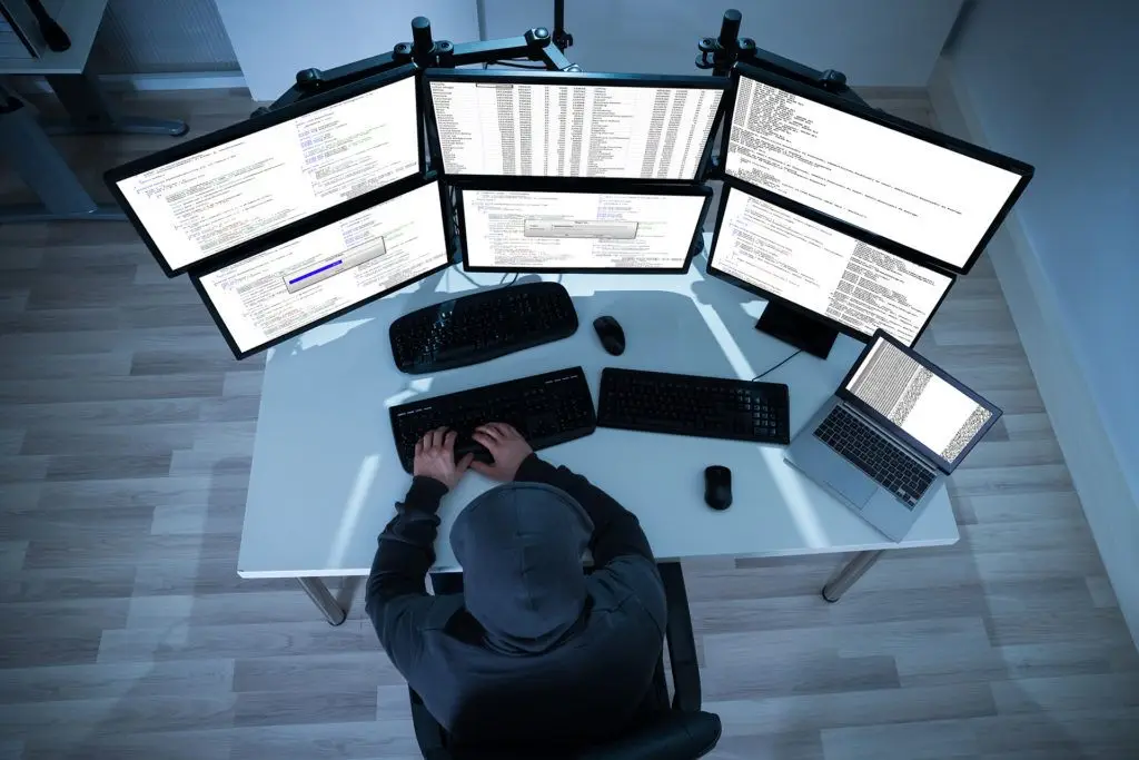 man in hoodie at a high-tech desk focused on cybersecurity programming across multiple screens.