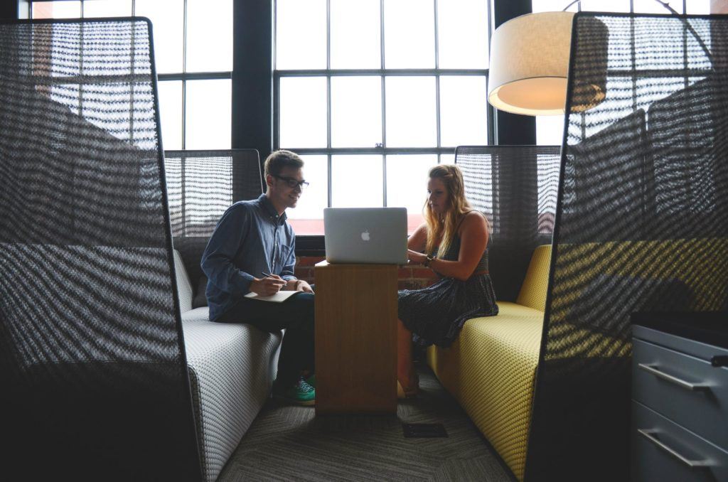 two professionals collaborate at a modern workspace table, engaging over a laptop in natural light.