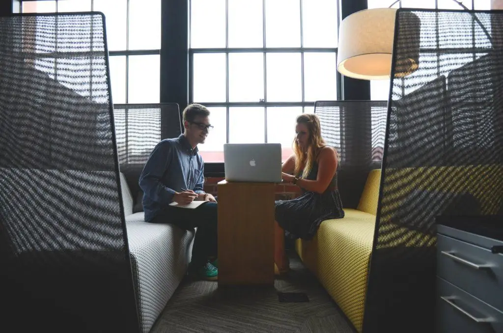 two professionals collaborate at a modern workspace table, engaging over a laptop in natural light.