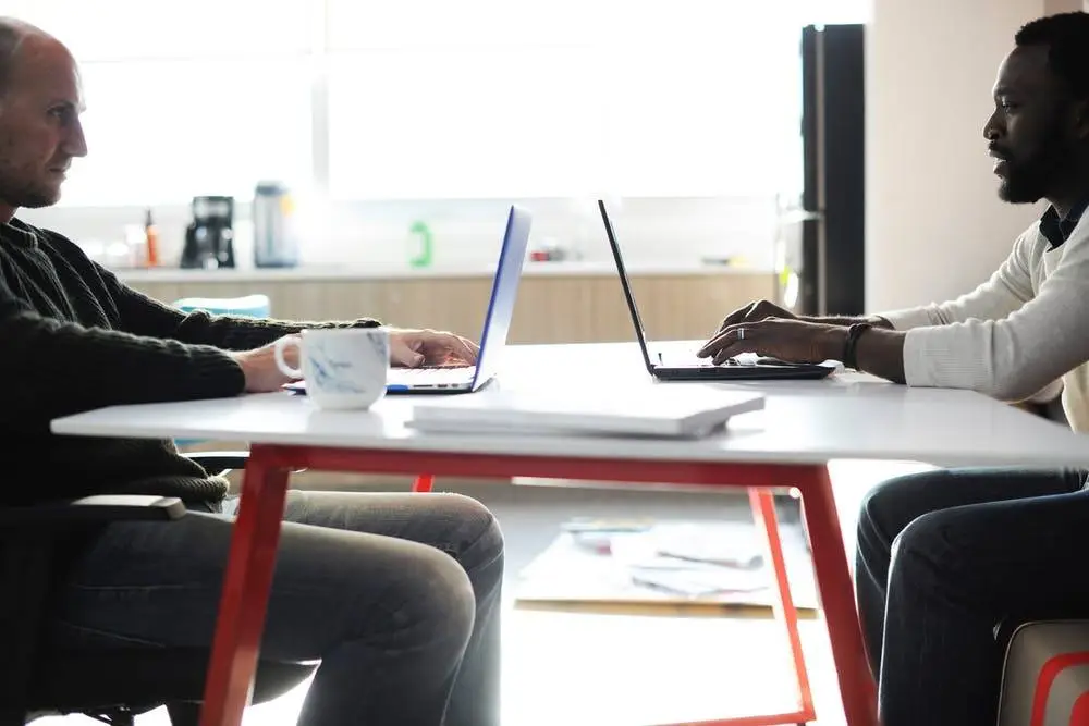 two professionals collaborate in a bright, modern workspace with laptops and a vibrant table.