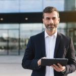 confident professional in a suit holding a tablet outside a modern corporate building.