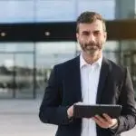 confident professional in a suit holding a tablet outside a modern corporate building.