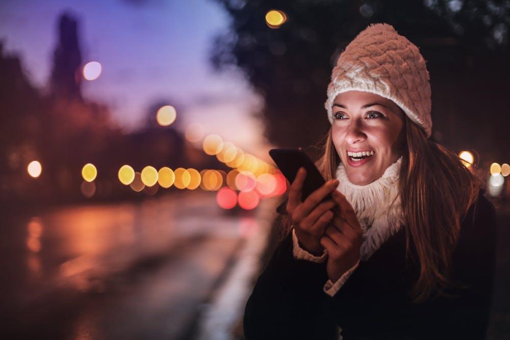 woman smiling at her smartphone outdoors in a warm, dusk cityscape, capturing a joyful moment.