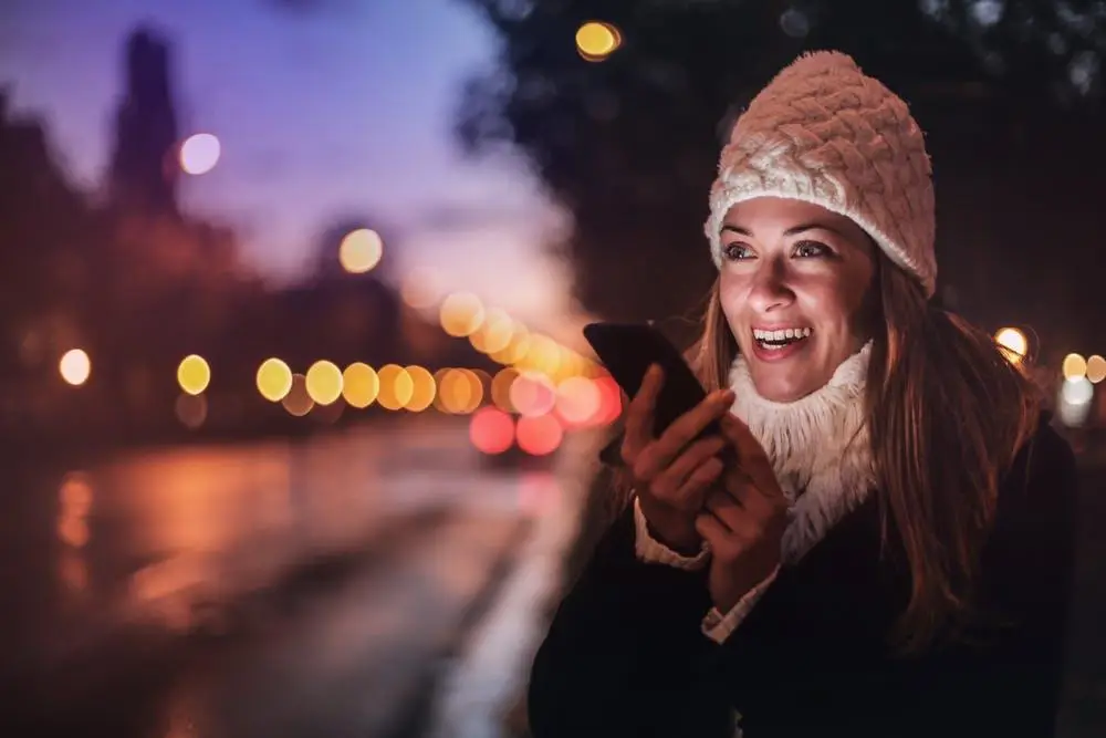 Voip Phone Services: What You Need To Know 1 woman smiling at her smartphone outdoors in a warm, dusk cityscape, capturing a joyful moment.