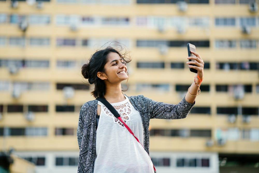 young woman joyfully taking a selfie in a sunny urban environment with flowing hair.