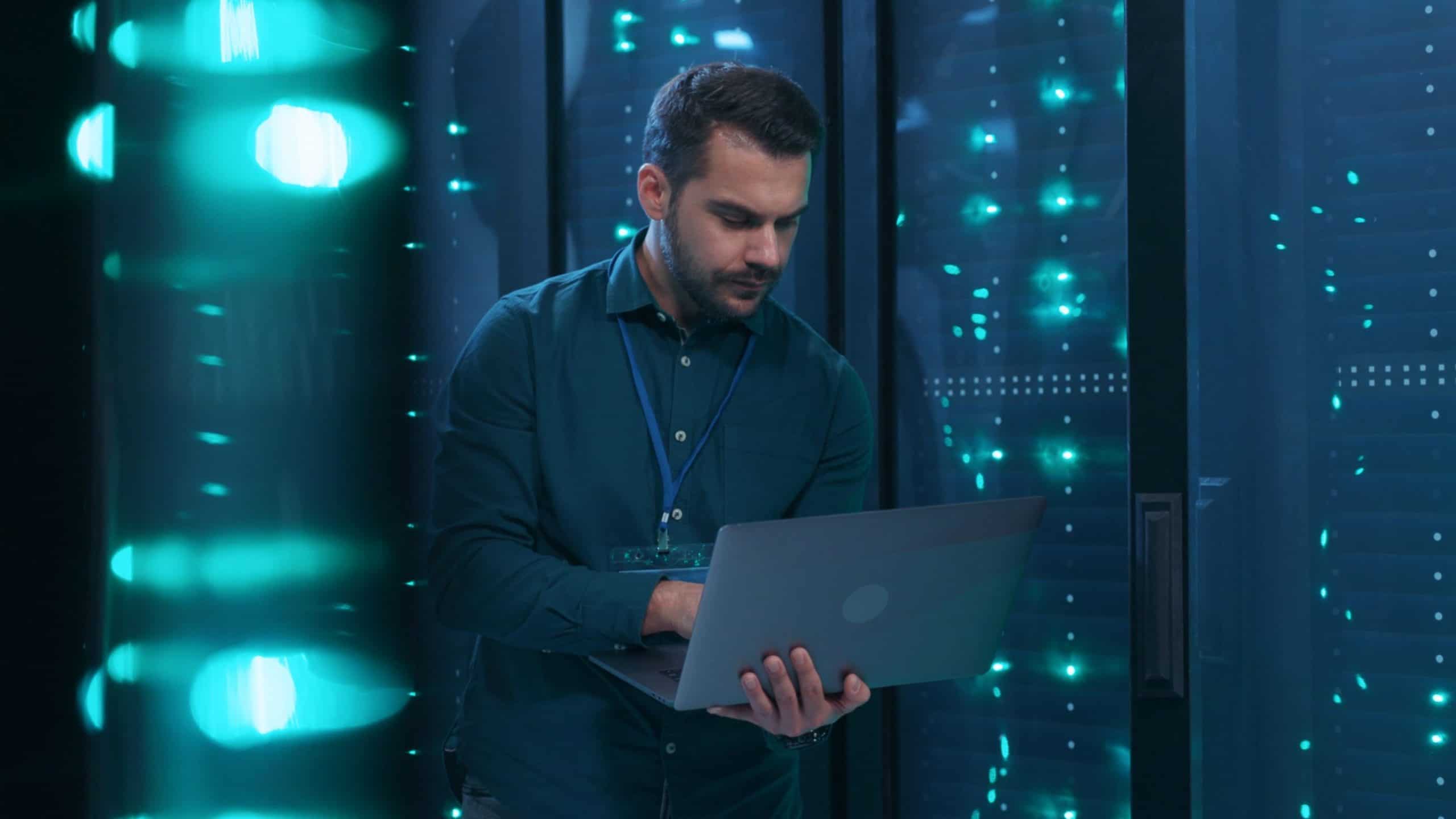 Caucasian IT professional admin using laptop computer doing data transfer operation with rack server cabinets in digital room of data center. Cyber security
