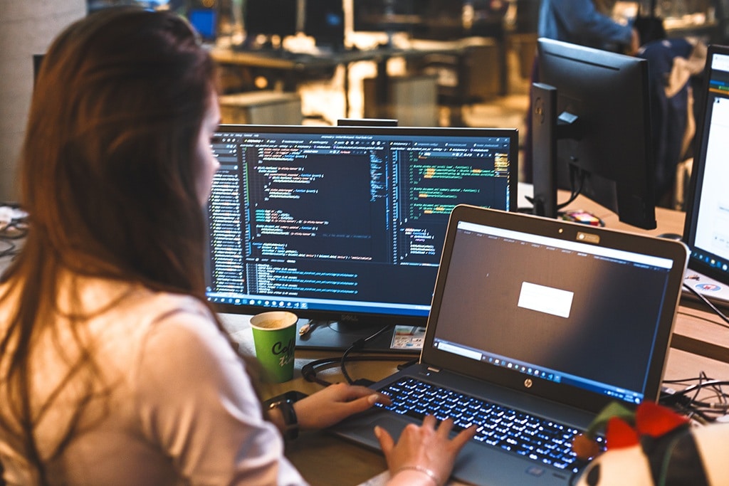 young woman coding on a laptop in a modern, minimalist tech workspace with dual monitors.