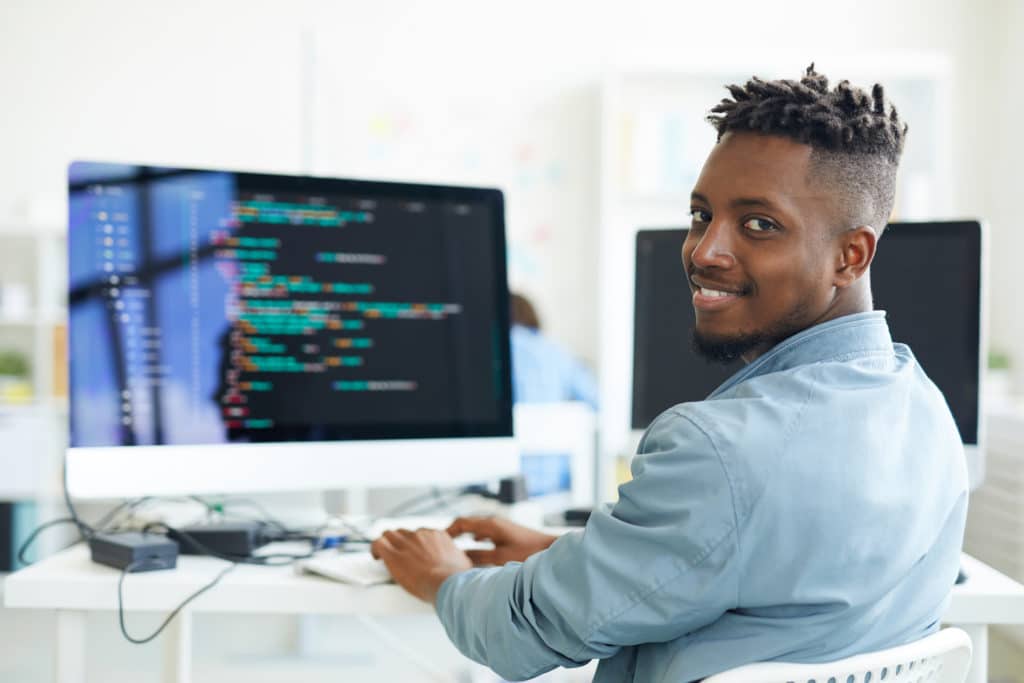 young programmer confidently coding at modern workspace with dual monitors and warm lighting.