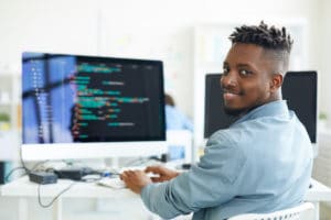 Happy guy looking at camera while sitting by workplace and working with software