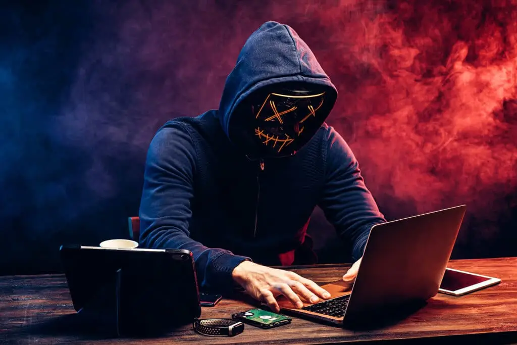 Differences Between Hollywood Hackers And The Real Deal 2 masked hacker at a table, illuminated by dramatic lighting, engaged in a secretive task.