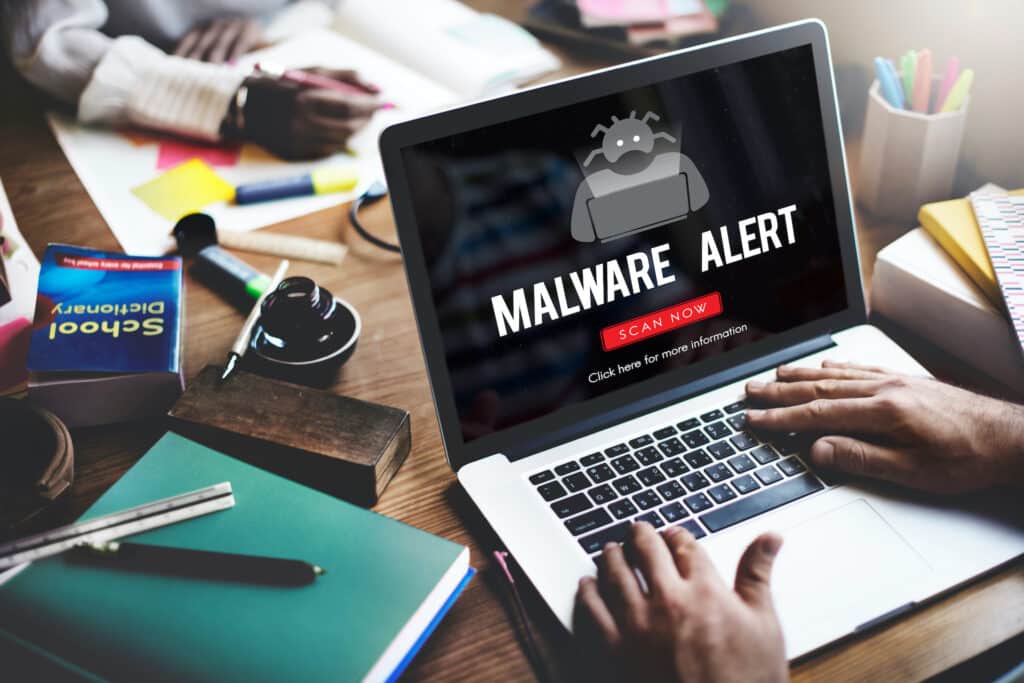 student facing a malware alert on a laptop amidst disordered study materials and textbooks.