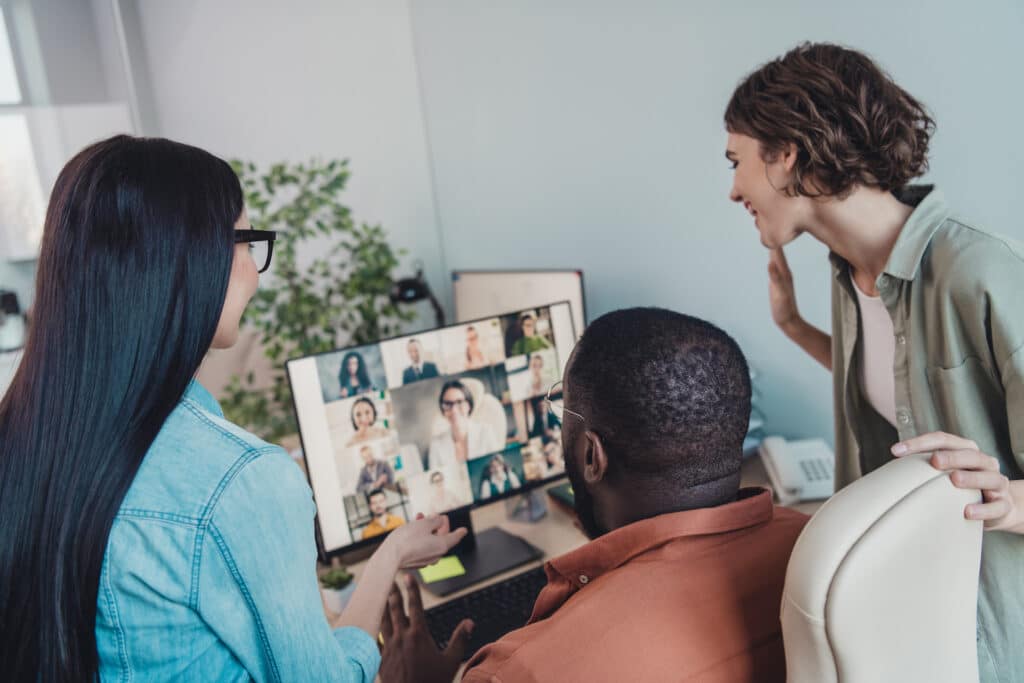 diverse team collaborates around a computer in a hybrid workspace, engaging in a video call.