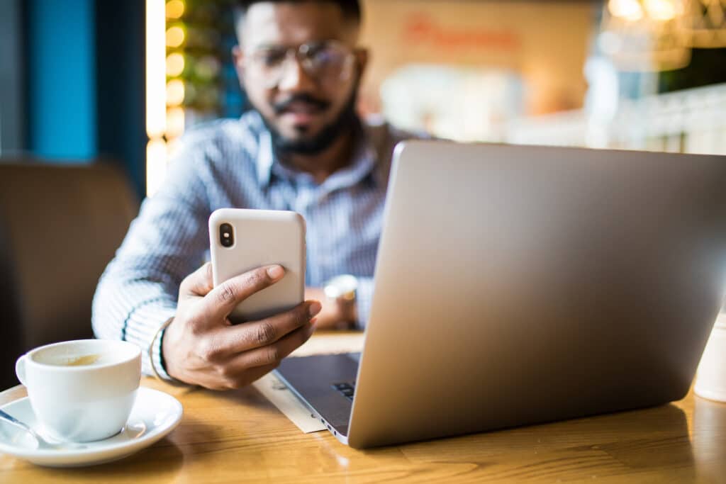 young indian professional typing on smartphone in cozy café with a laptop and coffee.
